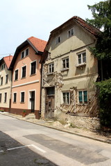 Fototapeta na wymiar Row of renovated attached old suburban family houses with one abandoned ruin at the end of street with dilapidated cracked walls and cracked wooden doors surrounded with paved road and tall trees