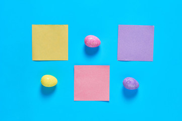 Three colorful eggs with glitters near blank square paper stickers lies on blue desk on kitchen. Easter concept. Space for text. Top view