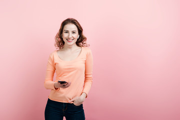 Portrait of smiling young woman holding mobile phone in hand, wearing wireless headphones, looking...