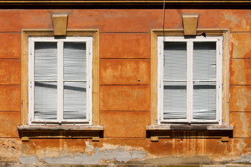 Two closed dilapidated windows with white wooden frames and broken plastic window blinds mounted on wall of old suburban family house with cracked facade