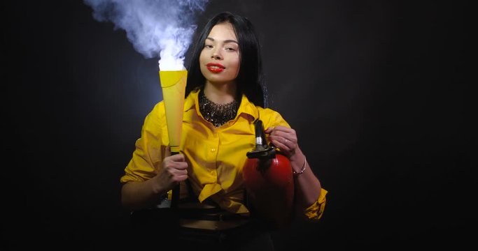 Funny and pretty brunette in a yellow shirt with a big red fire extinguisher from which steam comes on a black background isolated. Studio shot of surreal fashionable firefighter girl. 4k 