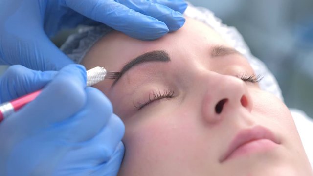 Beautician is painting shaping contour using manual tool for tattoo, woman's face closeup. Cosmetologist making eyebrows microblading procedure in beauty salon for girl. Beauty industry concept.
