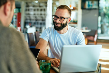 Attractive bearded man with glasses in a cafe working on laptop and chatting with a friend
