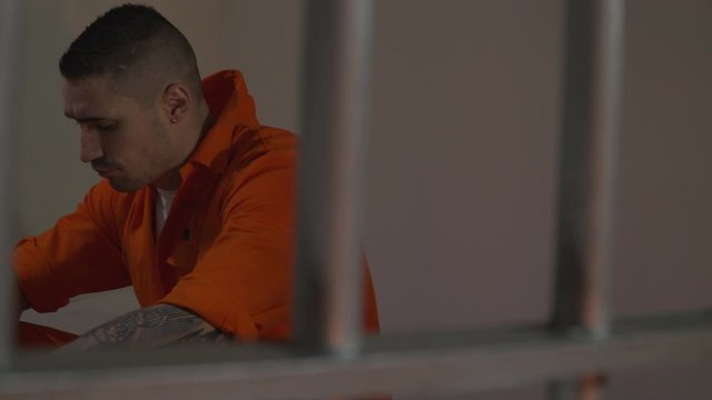 Male Prisoner behind bars in a Jail / Prison Cell. Close up. The criminal is sat on his bed, with his head down. Pan left.