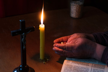 Wooden table in retro style. On it is a candle and a cross. Hands of an old woman over a book. Palms folded
