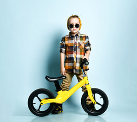 Positive boy in bright stylish casual clothing, sneakers and cap sitting on yellow run bike present...