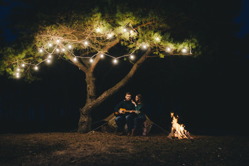 Young couple in love camping tourists sitting by a fire against a tent in the forest with a retro garland, photo with a lot of noise, selective focus