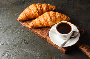 Espresso in a white cup and two croissants on a dark background, on wooden board, place for text, view from the top