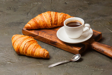 Espresso in white cup and two tasty croissants on a dark concrete background, on a wooden board. Concept for breakfast, coffee break or business lunch.
