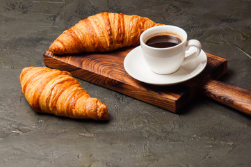 Coffee in white cup and croissant on a dark concrete background, on a wooden board. Concept for breakfast, coffee break or business lunch.