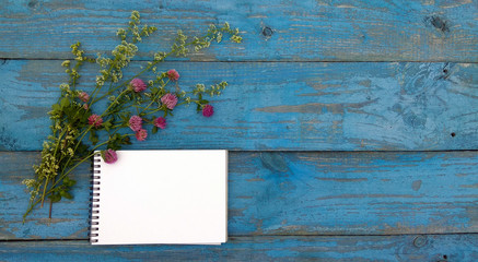 Obraz premium Mockup - notebook on wooden background. A sketchbook and a bouquet of clover lie on old blue boards. Flatlay for backgrounds, texts, desktops, banners, covers, postcards, to-do lists.