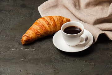 Croissant and cup of coffee and saucer on dark background with a gray linen napkin