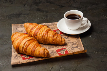 Croissant and cup of delicious coffee and saucer on craft paper on a dark background, vintage style, close up, top view