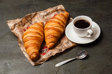 Delicious croissants on crumpled craft paper and espresso in a white cup with a spoon on dark gray background, vintage style