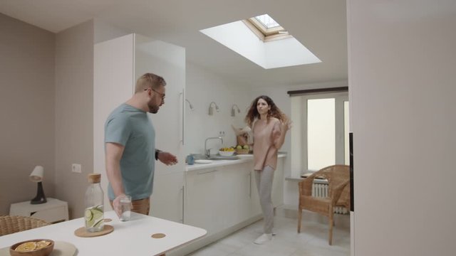 Dollying shot of young Caucasian woman washing dishes in kitchen, complaining and reproaching lazy boyfriend, then smashing plate and beating him with towel, and man throwing glass of water at her