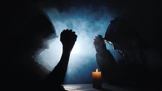 Two people sit at a table, praying. A candle burns near them, behind the mist refreshed by a spotlight