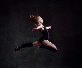 Young girl gymnast in black sport body and special uppers jumping in gymnastic pose over dark background