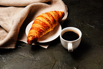Sweet croissant on saucer, a cup of ristretto coffee and a linen towel on a dark background