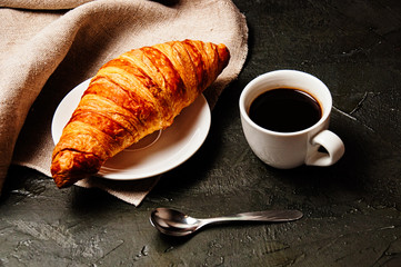 Sweet croissant on saucer, a cup of strong ristretto coffee, a spoon and a linen towel on a dark background