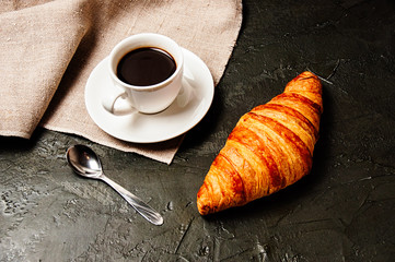 Sweet tasty croissant, cup of strong ristretto coffee on a saucer, a spoon and a linen towel on a dark background