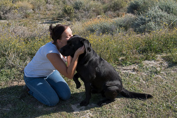 Woman sitting on ground cuddle and kiss Labrador dog while walking in nature.