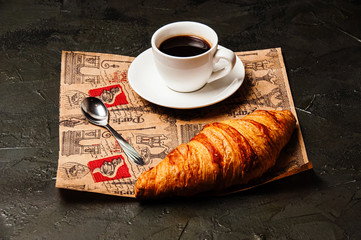 French croissant, sweet strong coffee in a white cup on a saucer with a spoon, on beautiful craft paper, on a dark background