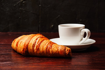 Croissant and coffee in white cup on dark wooden tabletop. Breakfast concept on the table