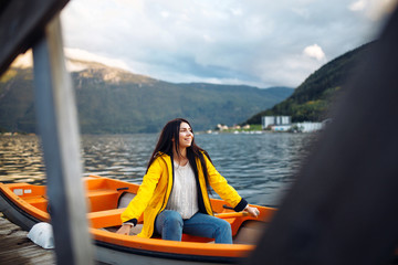 Fototapeta premium The girl tourist in a yellow jacket is sitting in a boat against the backdrop of the mountains in the Norway. Active woman relaxing on the boat by the lake. Travelling, lifestyle, adventure concept.