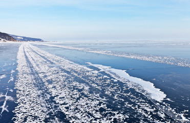 Baikal Lake on a cold day in January. An ice road along the coast to the village of Goloustnoye with snow tracks from the Hivus on the surface of blue ice. Winter landscape. Natural background
