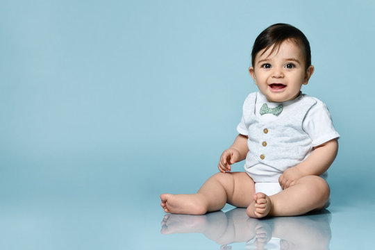 Baby Boy In White Bodysuit As A Vest With Bow-tie, Barefoot. He Smiling, Sitting On The Floor Against Blue Background. Close Up