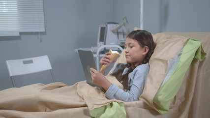 Girl lie on the bed, eat icecream and watch something on tablet