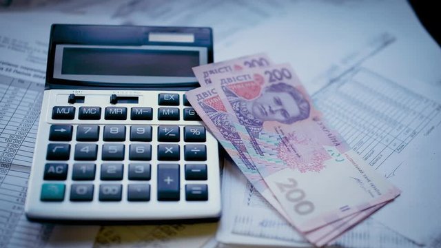 Next to the calculator, a few pieces of Ukrainian paper money worth 200 hryvnias fall on accounting documents. Slow motion.
