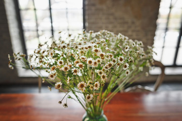 A charming bouquet of white little daisies in a glass jar on the wooden table.