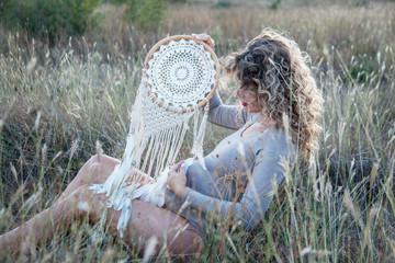 Young beautiful pregnant woman posing at the field of dried flower against the sunset. Girl wearing female pregnancy underwear and holding boho style dreamcatcher. Model soon to be mother