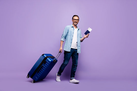 Full length photo of positive man hold big luggage tickets arrive abroad check-in like true plane airport passenger wear denim jeans shirt isolated over purple color background