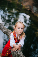 Little girl with white hair in a red, Slavic sundress. A girl walks in a birch forest by the pond