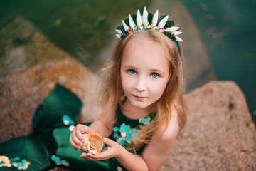 A little girl with white hair with a mermaid tail and shells is sitting on the shore. A little mermaid sits on a large stone by the pond.