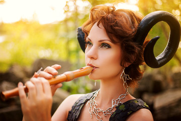 A girl in the image of a faun plays the wooden flute. Model with horns on his head and with furry pants in the woods among the stones - obrazy, fototapety, plakaty