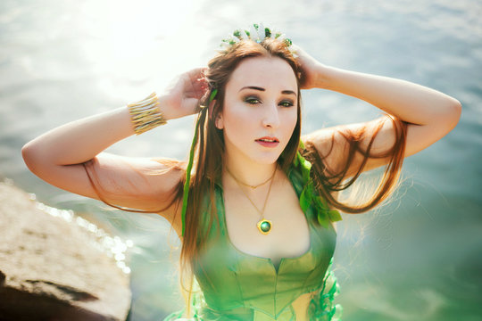 A girl with seaweed in her hair and in a green dress is standing near a pond. Model with a rim with shells. Mermaid near the river
