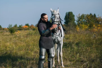Tuinposter A knight strokes a white horse. Armored guy with a white horse. A man in chain mail. Image of a warrior © Вероника Преображенс