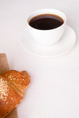 croissant in a paper box and coffee in a white cup on a white table