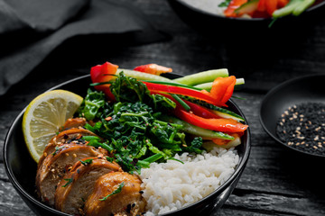 Two lunch bowls with teriyaki chicken, cucumber, red bell pepper, green onion, stew spinach and basmati rice on a rustic black table.