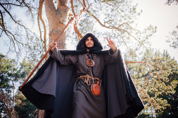 A man with a beard and a mustache in a robe and coat with a staff in his hand. A magician, a shaman conjures in the forest. Fantasy image