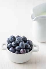 Close-up of blueberries in white bowl, on white background, with white milkmaid in the background out of focus and copy space