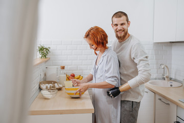 Wife cooks, whisking mixture in a bowl, while her husbang stays behind