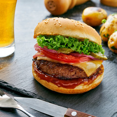 Cheeseburger made of meat burger, cheese, tomato, lettuce, onion and pickled cucumber with baked potato and broccoli served with glass of beer and sauce.