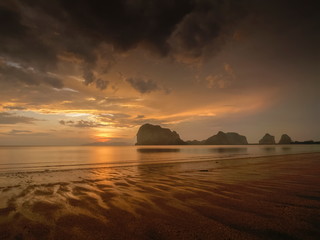 view seaside evening of mountains on sand beach with yellow sun light and dark clouds moving in rainy sky background, sunset and raining at Pak Meng Beach, Trang Province, Thailand.