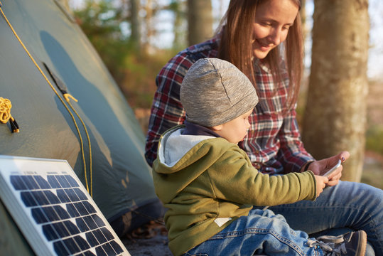 Little Wonderful Kid Takes The Phone From A Mother Sitting Near The Solar Panel At A Campsite In The Forest Under The Sun's Rays. Child Dressed In Jeans, Jacket And Hat In The Middle Of Warm Autumn