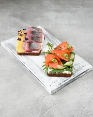 Two smorrebrod with herring, trout and vegetables on white wooden board