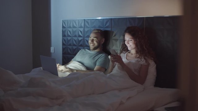 Full shot of young Caucasian couple of millennials in pyjamas sitting up in bed at night, leaning on headboard, using laptop and smartphone, smiling, laughing and showing something to each other